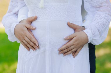 Nazikçe sarmaş dolaş hamile bir karın, sevgi, beklenti ve beyaz elbiseli bir kadın ve bir ortağın desteğiyle yeni bir hayat beklemenin sevinci..