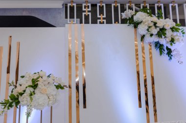 Beyaz bir duvar içeren zarif bir düğün arkaplanı. Dikey altın metal barlar ve çiçeklerle süslenmiş beyaz güller ve yeşillik etkinliği dekorasyonu..