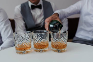 Sofistike giyinmiş bir adam tarafından üç bardak kehribar viskisi dolduruluyor. Muhtemelen bir kutlama ya da karanlık bir geçmişi olan bir tost..