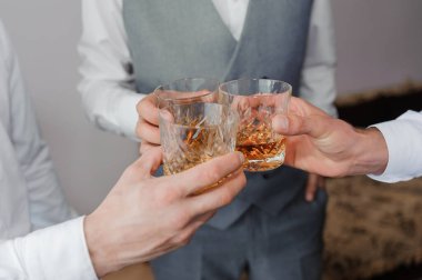 Kutlamada bardakları kehribar viskiyle dolduran iki adam, muhtemelen bir düğünde ya da resmi bir etkinlikte, gri yelek ve beyaz gömlekler giyerek..