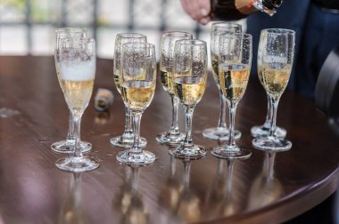 Bir sıra zarif dolu şampanya flütleri, kabarcıklarla parıldıyor, parlak, koyu ahşap yuvarlak bir masanın üzerinde duruyor, yakınlarda bir şişe, bulanık bir arka planla kutlama kadeh kaldırmayı öneriyor..