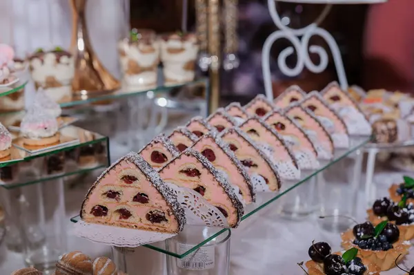 Kiraz dilimleri, küçük kekler ve çikolatalar içeren bir dizi hamur işi sergiliyor. Cam tabaklar üzerinde çiçek aksanıyla sunuluyor.