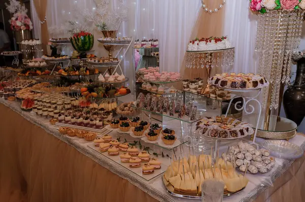 Bol şekerli ve dekorasyonlu savurgan düğün masası görüntüsü Altın masa örtüsü beyaz perdeler kristal avizeler zarif karşılama ayarı.