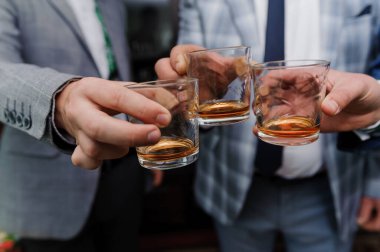 Yakın çekim üç adamı, muhtemelen meslektaşlarını ya da arkadaşlarını, kadeh kaldırmak için viski bardaklarını tutarken yakalar. Adamlar takım elbise ve resmi giyinmişlerdir.