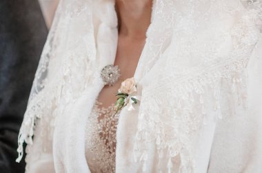 Detaylı bir çekim, narin beyaz dantelli bir düğün ceketi giyen gelinin göğsünün üst kısmına odaklanır, parlak bir broş ve tek bir allık gülü ile vurgulanır.