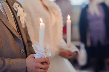 Düğün mumu seremonisi sırasında damat ve gelinin yakın çekim sahnesi, ellerinde beyaz mumlar, geleneksel kıyafetler ve resmi ifadeler vardı.
