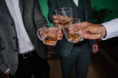 Yakın plan çekimde, 30 'lu ya da 40' lı yaşlarda koyu takım elbiseli, loş, ince bir atmosferde kehribar renkli viski kadehleri kaldıran üç adam resmedilmiş.