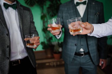 Bir grup akıllı giyimli sağdıç kadehlerini çağdaş bir mekanda, yemyeşil yapraklarla vurgulanmış bir şekilde kaldırarak bir düğün önerisinde bulunuyorlar.