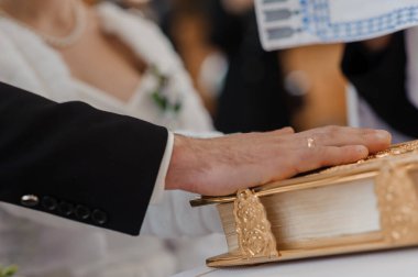 Gelin ve katılımcıların bulanık arkaplanda kutsal bir yemini sembolize eden, güzel dekore edilmiş altın bir kitabın üzerinde duran damadın eline odaklanan bir düğün töreninin yakın çekimi.