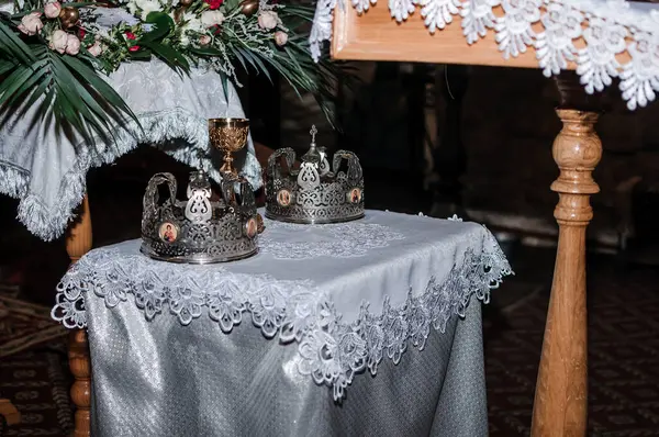 Sergilenen dinsel bir sahne, gümüş taçlar, altın bir kadeh, ve bir kilisenin karanlığında karışık dantellerle bezeli bir masa üzerine kurulmuş güzel bir çiçek aranjmanı.