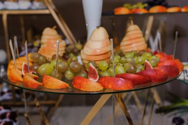 Dilimlenmiş armutlar, incirler ve yeşil üzümlerden oluşan lüks bir meyve tabağı cam tezgahta zarif bir şekilde yerleştirilmiş, lüks etkinlikler, düğünler veya yemek servisi için mükemmel, zengin, renkli ve zarif bir sunum sunumu olan