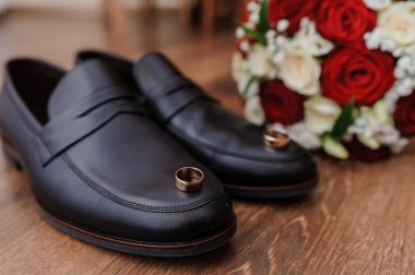 Ilık ahşap bir masa üzerine serpiştirilmiş, yemyeşil bir buket kırmızı ve beyaz gül eşliğinde, parlak siyah deri ayakkabılar üzerinde dinlenen iki altın nikah yüzüğünün yakın plan çekimi.