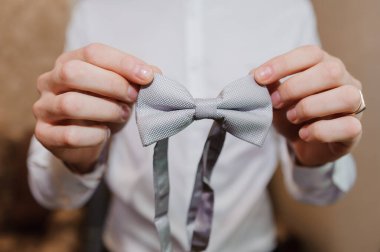 Yakın çekim, bir damadın ellerini açık gri bir papyonu dikkatlice tutarken gösterir. Düğüne hazırlanırken, resmi hazırlıkları ve kutlamaları önerir.