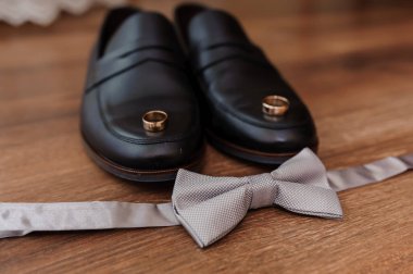 Zarif bir düğün hazırlığı, siyah deri ayakkabılar, açık gri bir papyon ve sıcak ahşap bir masada serili altın alyanslar, lüks ve romantizm dolu.