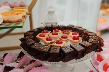 Çürümüş çikolatalı vişneli kek taze kirazlarla süslenir. Pembe ve diğer çeşitli pastalarla çevrili beyaz bir pasta standı. Fırın vitrininde güzelce sunulur.