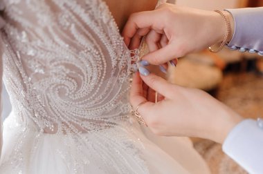 Hassas bir an, narin dantel detaylar ve yumuşak lavanta aksanlarıyla karışık boncuklu gelinliğiyle bir gelinin sırtını gösteriyor.