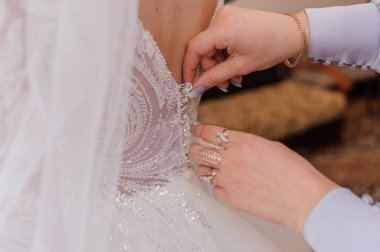 Gelinin sırtına sıkıca çekilen bir gelinliğin yakın plan çekimi. Karmaşık danteller ve inciler sergileniyor. Sıcak pembe renkler ve yumuşak bir odak romantik ve zarif bir ortam yaratıyor.