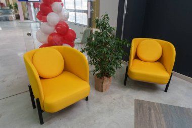 Parlak bir ofis lobisinde büyük bir pencerenin yanına yerleştirilmiş iki rahat sarı koltuk ve dekoratif kırmızı bir balon düzenlemesi yer alıyor.