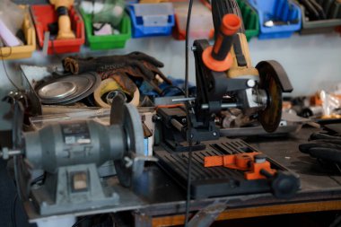 Dağınık bir atölye tezgahında gri bir arkaplan üzerinde eldiven ve çeşitli parçalar içeren turuncu bir bank öğütücüsü sergileniyor.