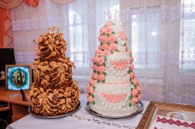 Bir çift özenle hazırlanmış, pembe rozetlerle süslenmiş beyaz düğün pastası çerçevelenmiş portrelerin yanında duruyor, rahat, klasik tarzda bir evde kutlama sahnesi sergiliyor.