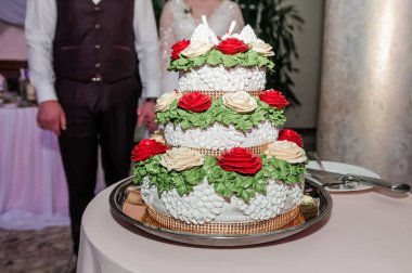 Üç katlı güzel bir düğün pastası odak noktasıdır, bol bol beyaz şekerleme, canlı kırmızı güller ve taze yeşil yapraklarla süslenir, bir festival resepsiyonunda sergilenir.