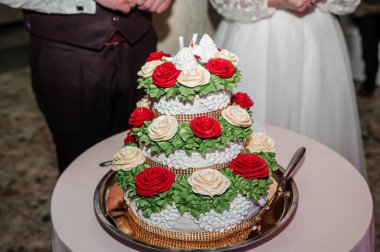 Kırmızı güller ve yemyeşil çiçeklerle süslenmiş güzel dekore edilmiş üç katlı beyaz bir düğün pastası zarif bir balo salonunda düzenlenen düğün kesim töreninde sahneye çıkıyor.