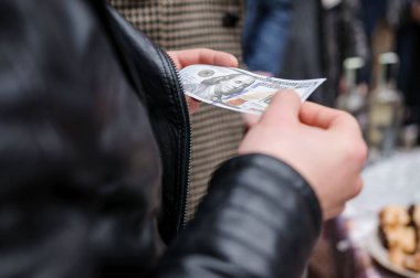 Yakın plan bir para alışverişini tasvir ediyor, siyah deri ceket giyen birine 100 'lük veren bir ele odaklanmak sokak seviyesinde bir alışveriş izlenimi uyandırıyor, bulanık bir arka plan, gizlilik hissi katıyor.