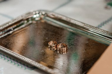 Yakın plan çekimde iki altın alyans, karmaşık kabartmalı detaylarla birlikte gümüş bir tepside duruyor. Aşkı, bağlılığı ve evliliği simgeliyor.