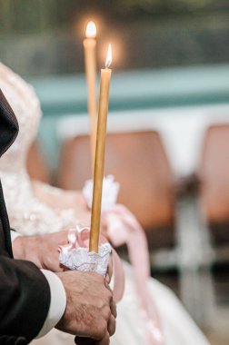 Ortodoks düğün mum yakma seremonisini yakalayan güzel bir kare, gelin ve damadın, yumuşak şeftali ve altın tonlarıyla kilisenin içinde yanan konikleri tutarken, sıcak ve geleneksel bir atmosfer yarattığını gösteriyor.