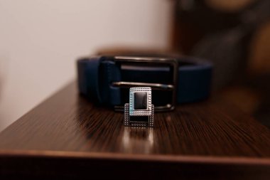 Yakın plan çekimde parlak mavi bir deri kemer ve zengin ahşap bir yüzey üzerinde duran gümüş toka, düğün hazırlıklarını ve erkeklerin resmi tarzını sembolize ediyor.