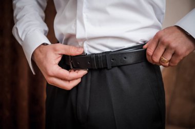 Siyah takım pantolonun üzerine klasik bir siyah deri kemer takan, beyaz bir gömlek giyen ve tımar etmeyi, resmi giyinmeyi, stili ve lüksü öneren altın bir yüzük takan bir adam.