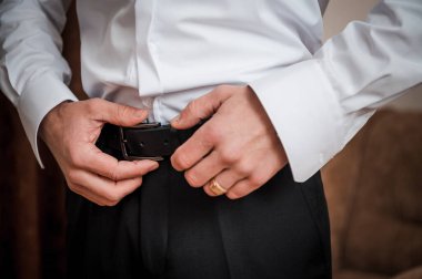 Yakın çekim, beyaz gömlekli ve siyah pantolonlu bir adamı gösteriyor. Deri kemer takıyor, altın bir alyans takıyor, düğün hazırlıkları sırasında, zarafet öneriyor.