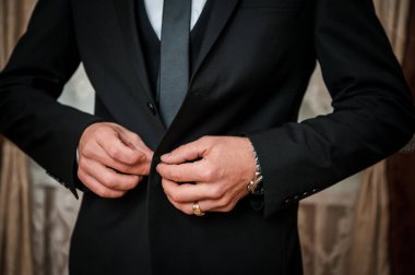 Klasik siyah smokinli yakışıklı bir adam resmi bir etkinlikten önce kol düğmelerini ayarlıyor, muhtemelen bir düğün, lüks bir saat ve zarif bir stil sergiliyor.