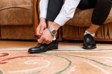 Bir damat, kol saatini ayarlarken siyah deri bağcıklı ayakkabılarını dikkatlice bağlar ve lüks bir iç mimariye sahip sofistike bir düğün günü hazırlık sahnesini gözler önüne serer.