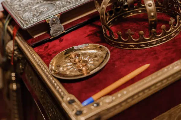 Çarpıcı bir natürmort altın bir taç, tabakta alyanslar, antika bir tüy kalem ve koyu kırmızı kadife bir yüzey üzerinde dinlenen zengin süslemeli bir el yazması tasvir eder.