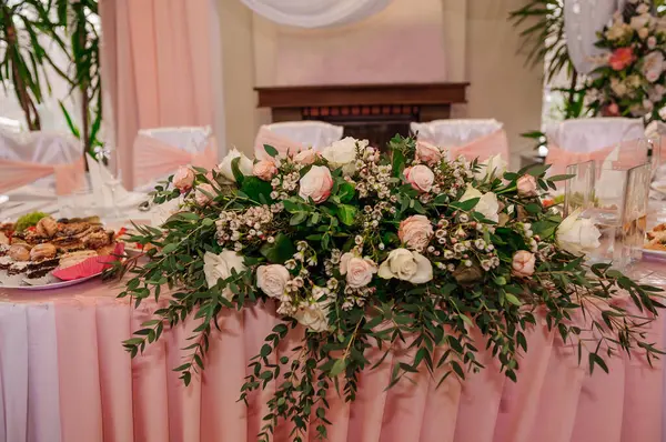 Müsrif bir düğün resepsiyonu, pembe güller, ortancalar ve yumuşak pembe tekstil örtüleriyle süslenmiş çiçeklerle süslenmiş uzun bir masa sergiliyor.