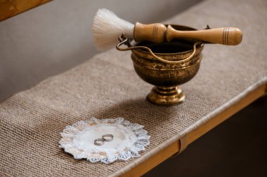 Yakın plan çekimde dantelli çarşafların üzerinde sunulan alyansların yanında pirinç tütsü yakıcı ve fırça, yumuşak ve sıcak tonlarda gelenek ve samimiyeti çağrıştırıyor.