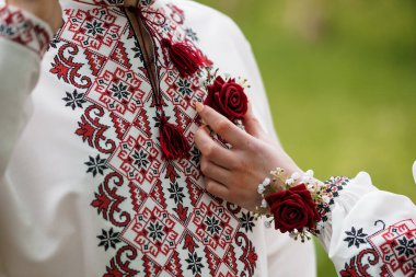 Güzel işlemeli beyaz Ukrayna vyshyvanka tişörtü, canlı kırmızı çiçek desenleriyle süslenmiş ve kırmızı güllerle bir kadının elinden tutularak geleneksel kültür mirası ve halk sanatı tasvir edilmiş.
