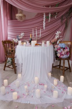 Büyüleyici romantik bir yemek sahnesinde beyaz çarşaflar, mumlar ve narin çiçeklerle süslenmiş bir masa var. Yumuşak pembe bir arka plana karşı.