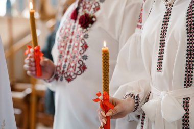 Geleneksel Ukrayna Ortodoks düğün töreninde balmumu mumlarını tutan ellerin yakın çekimi kırmızı aksanlı beyaz işlemeli gömlekler maneviyatı ve kutlamayı temsil ediyor.