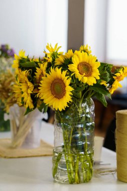 Beyaz bir masanın üzerinde duran şeffaf bir plastik şişe vazoda sergilenmiş canlı bir ayçiçeği buketinin yer aldığı güzel bir yaşam kompozisyonu. Güneşli sarı çiçekler ve doğal ışık neşeli ve davetkar bir sahne yaratır.