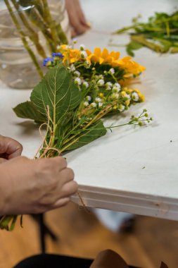 Yakın plan çekimde, küçük bir buket canlı sarı ayçiçeği, narin beyaz bebek nefesi ve yemyeşil yapraklar bağlayan, çiçek düzenleme atölyesini resmeden eller görülüyor.