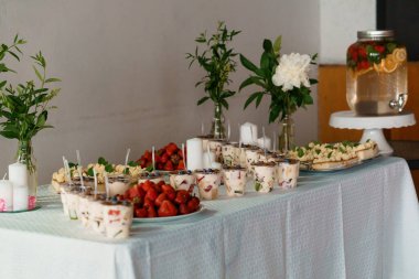 Çok güzel düzenlenmiş bir tatlı masasında çeşitli çilek parçaları, çeşitli pastalar, taze çilekler ve çiçekli süslemelerle tamamlanmış narenciyeli bir su dağıtıcısı bulunur.