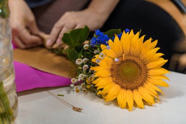Eller, sıcak tonlu bir atölyede narin mavi Delphinium ve papatya çiçekleri ile canlı bir ayçiçeği buketi düzenliyor. Huzurlu ve yaratıcı bir atmosferi iletiyor.