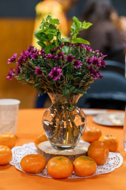 Güzel, durgun bir yaşam düzeninde, turuncu masa örtüsüne dağılmış portakallı cam bir vazoda mor kasımpatılar sergilenir.