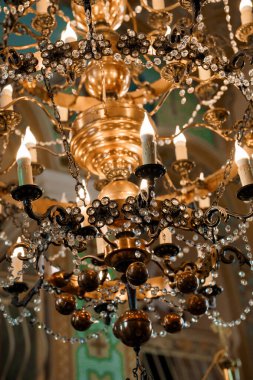 Yakından bakınca, pırıl pırıl kristal damlacıklarla süslenmiş muhteşem bir antika avize sergileniyor. Ilık mum ışığıyla aydınlatılmış, yaldızlı bir iç mekânda zengin bir barok lüksüne sahip.