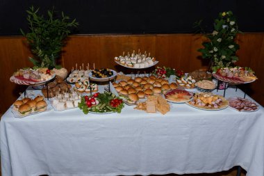 Bu resim, çeşitli et, peynir, mini sandviç ve canlı çiçeklerle dolu uzun beyaz bir masa sergiliyor. Etkinlik düzenlemek için ideal.