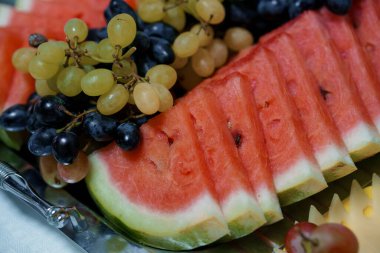 Göz alıcı bir yakın çekim natürmortu, yeşil ve siyah üzüm kümelerinin yanında sulu karpuz dilimleri içeriyor. Bu da ferahlatıcı bir yaz atıştırması veya meyve salatası hazırlığı öneriyor.