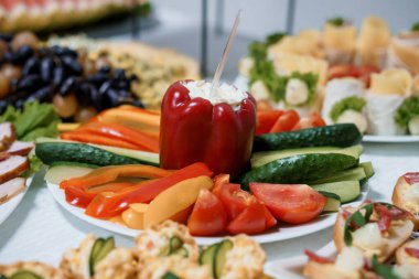 Renkli bir aperatif, havuçlar, salatalıklar, domatesler, zeytinler ve kremalı soslar ile çevrili dolgulu kırmızı bir çan biberi sergiliyor. Parti yemekleri ve sağlıklı atıştırmalıklar için ideal.