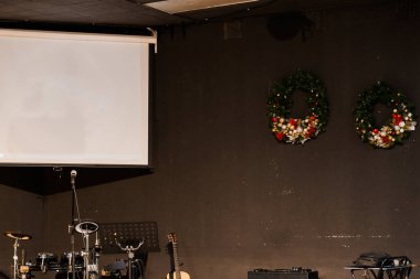 Noel için boş bir sahne, boş bir projeksiyon ekranının altında kahverengi bir duvarı süsleyen iki çelenkle süslenir, içinde davul seti, klavye ve müzik aletleri bulunur.